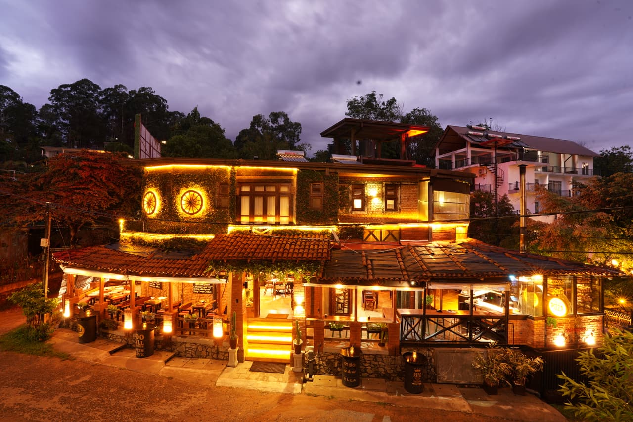 AK Ristoro on Passara Road, Ella, at blue hour — every floor lit, the steps glowing and two lit wagon wheels under a stormy sky